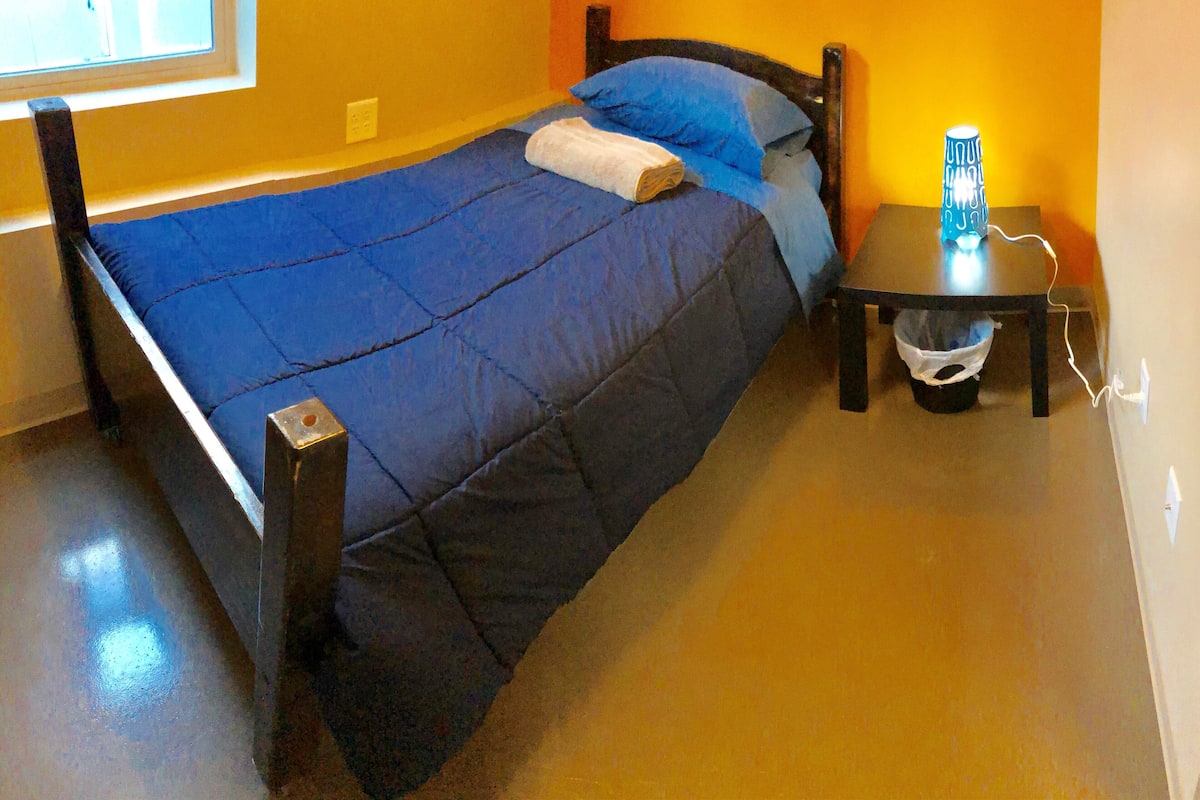 A bed with a blue and black bedspread, a wooden headboard, and a bedside table with a lamp.