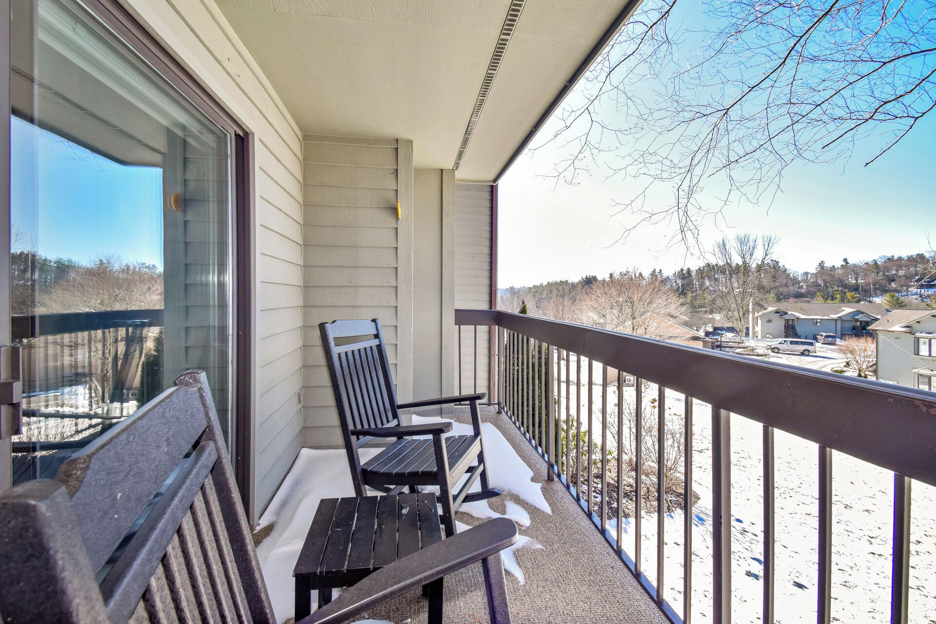 Un balcon avec un banc en bois et une chaise, donnant sur un paysage enneigé parsemé de maisons et d’arbres.