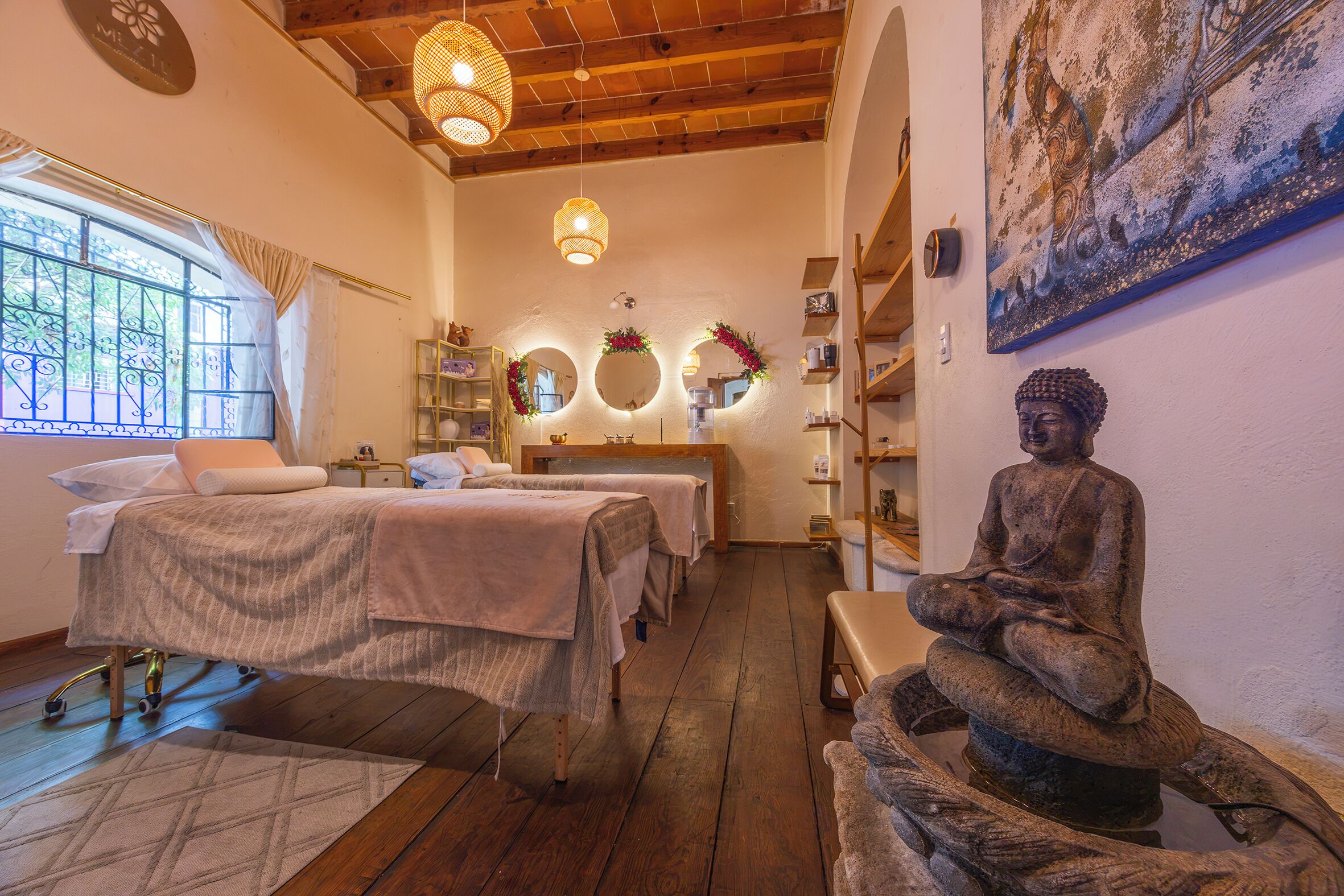 Un cuarto de spa con pisos de madera, dos camas, un rincón de meditación con una estatua y un amplio ventanal con detalles de hierro forjado.