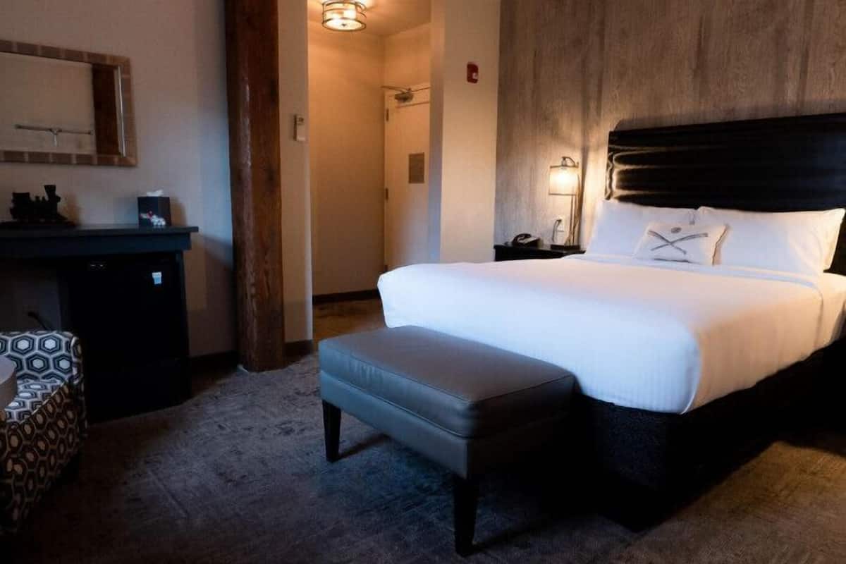 Habitación de hotel con una cama grande, una mesita de noche con lámpara, un escritorio pequeño y una silla estampada.