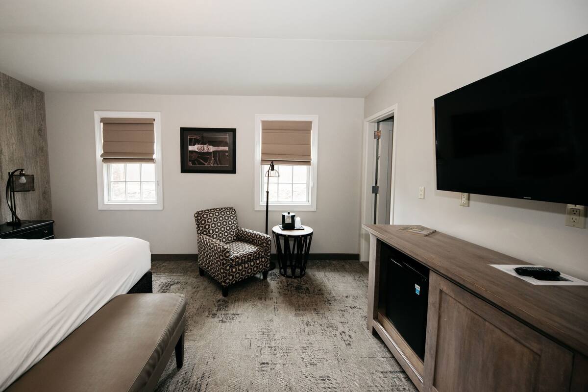 Una habitación de hotel con cama, zona de estar con una silla y una mesita, televisor y mueble de madera.