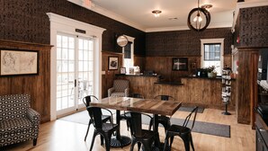 A dining area with a wooden table and chairs, a patterned armchair, and a kitchen area with a wooden counter and cabinets. - The Railroad Inn (Cooperstown)