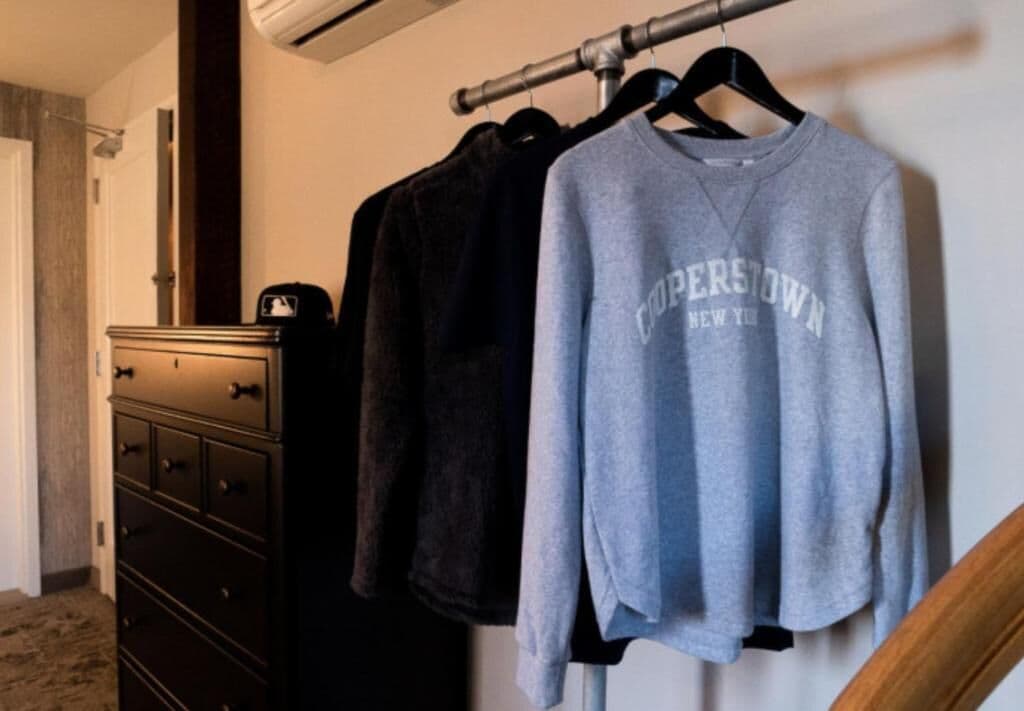 A room with a dresser, a hanging rack with clothes, and a grey sweatshirt with 'COPENHAGEN NEW YORK' text.