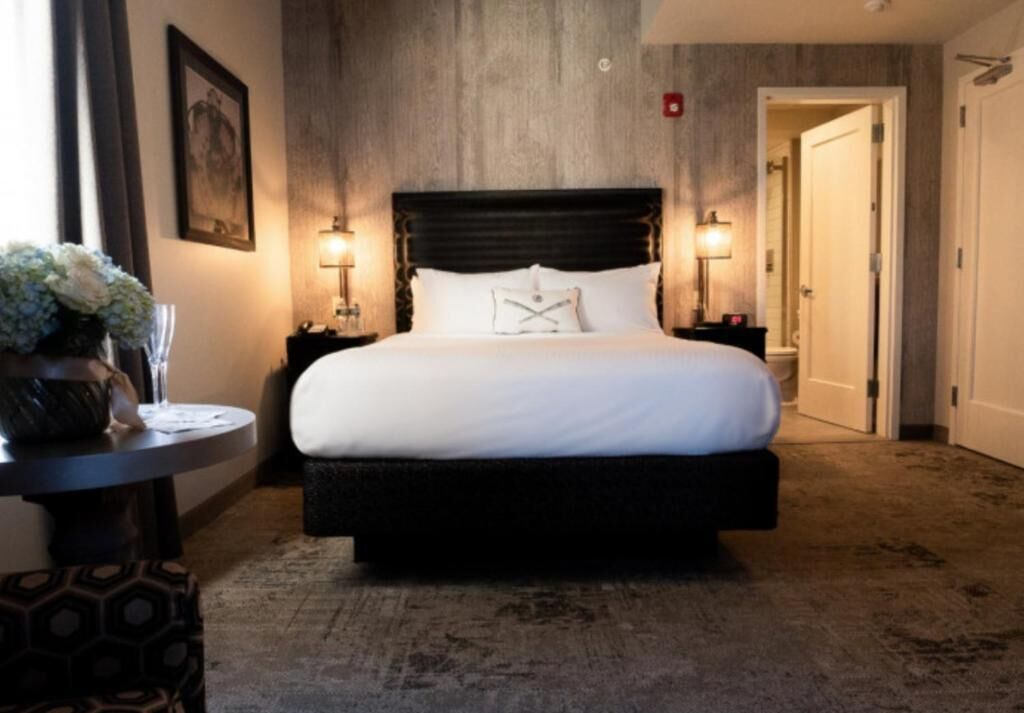 Een hotelkamer met een groot bed, twee bedlampen, een ronde tafel met een vaas, en een deur naar een andere kamer.