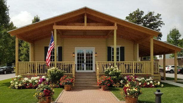 A wooden building with a porch, a flag, and flower beds. - The Railroad Inn (Cooperstown)