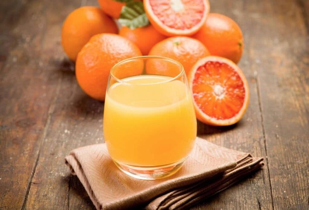 A glass of orange juice on a wooden surface with oranges in the background.