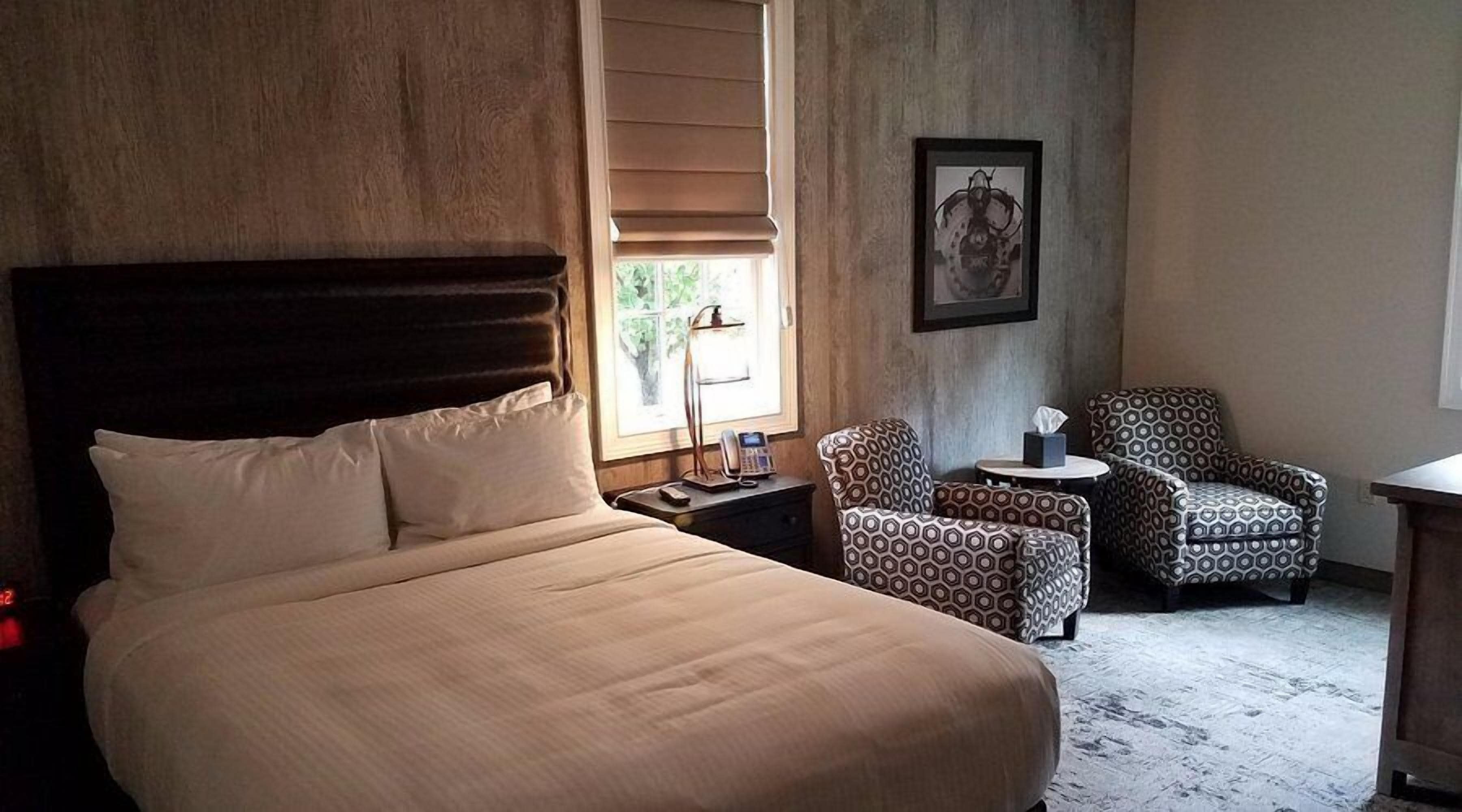 A hotel room with a large bed, two patterned armchairs, a small table, and a framed picture on the wall.