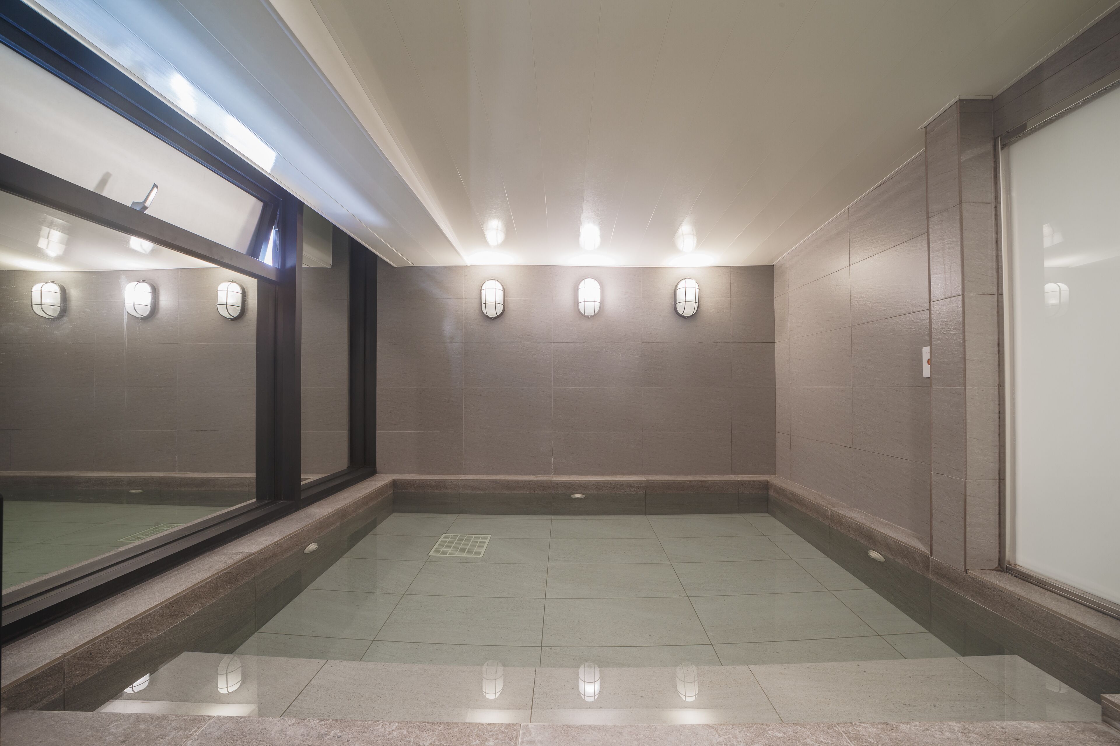 A spacious, well-lit indoor area with tiled floors and walls, featuring a large window on one side and wall-mounted lighting.