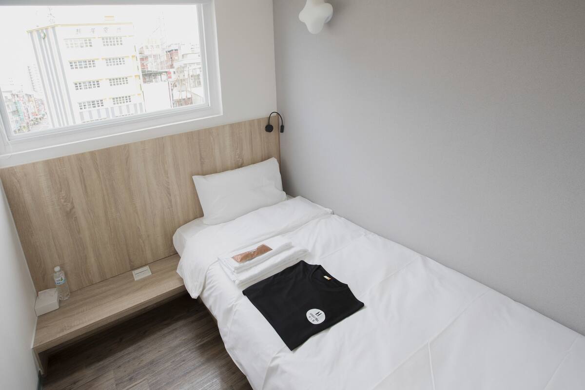 Ein kleines, ordentlich bezogenes Bett mit weißer Bettwäsche, einem Holzkopfstück und einem Fenster mit Blick auf Gebäude.