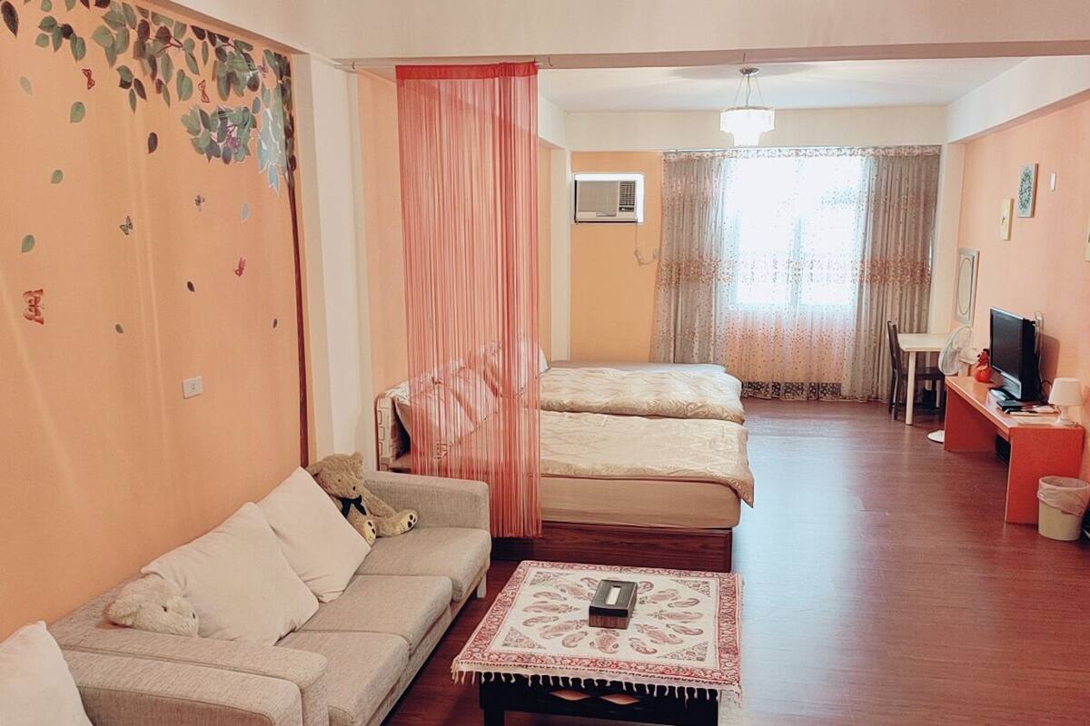 A hotel room with a sofa, two beds, a small table with a book, a TV, and a window with curtains.