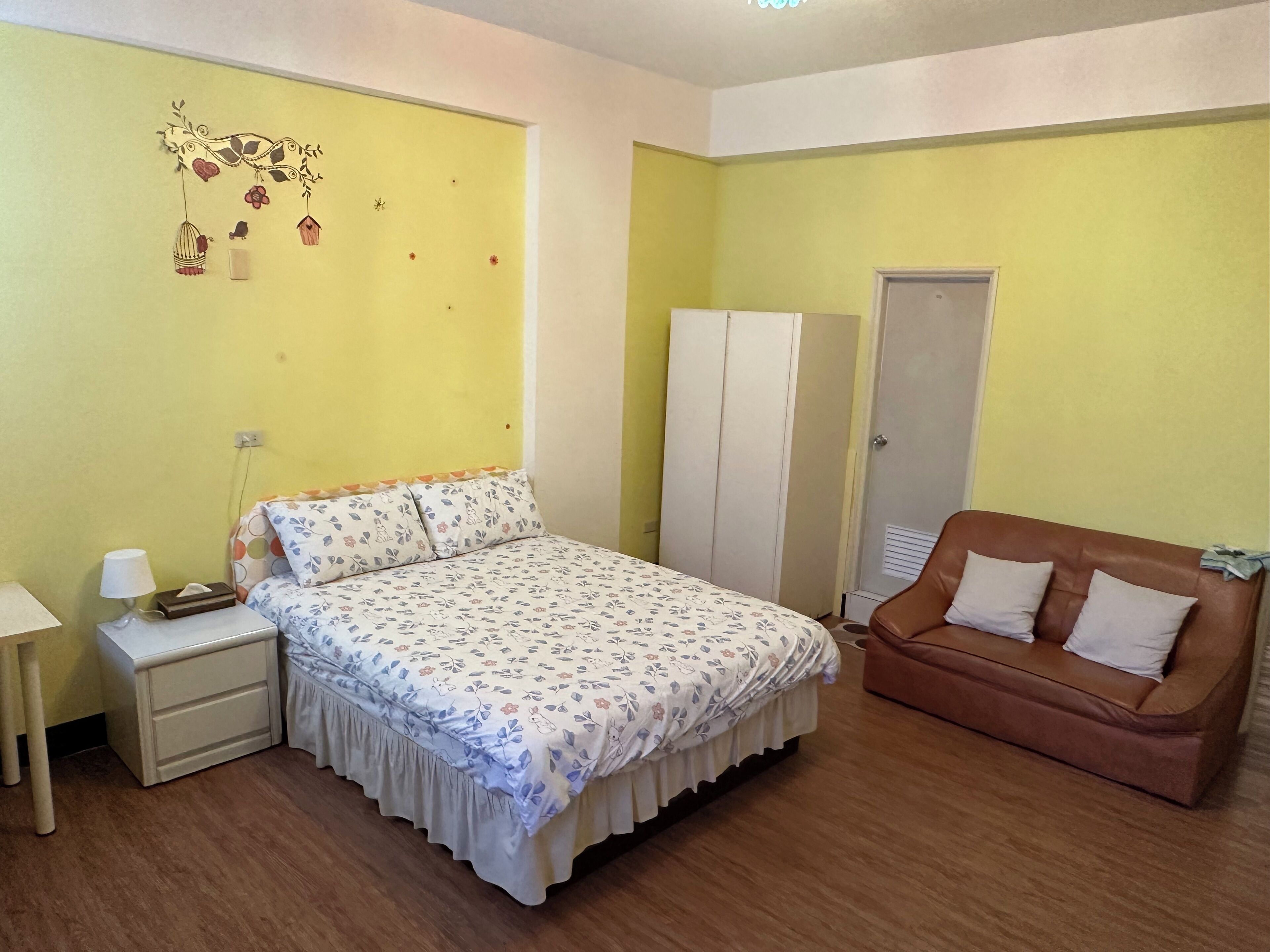 A bedroom with a bed, a nightstand, a lamp, a wardrobe, and a brown sofa.