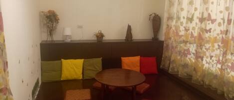 A cozy living room with a round wooden table surrounded by colorful cushions, a sofa with patterned cushions, a decorative vase, and a wall-mounted lamp.