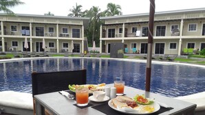 Une table à manger, dressée avec des aliments et des boissons pour le petit-déjeuner, est placée au bord d’une piscine, d’où l’on peut admirer un hôtel doté de nombreux balcons et bordé de palmiers.