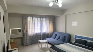 A bedroom with a bed, a small table, a blue sofa, a window with curtains, an air conditioning unit, and a ceiling fan. - Tainan Here Hostel 2 (Tainan)