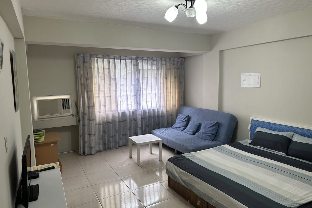 Quarto com cama, mesinha, sofá azul, janela com cortinas, ar condicionado e ventilador de teto.