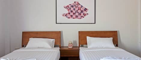 Ein Hotelzimmer mit zwei Einzelbetten, Holz-Kopfteilen, weißen Laken, einem kleinen Nachttisch mit einem Teddybären und einem gerahmten Kunstwerk an der Wand.