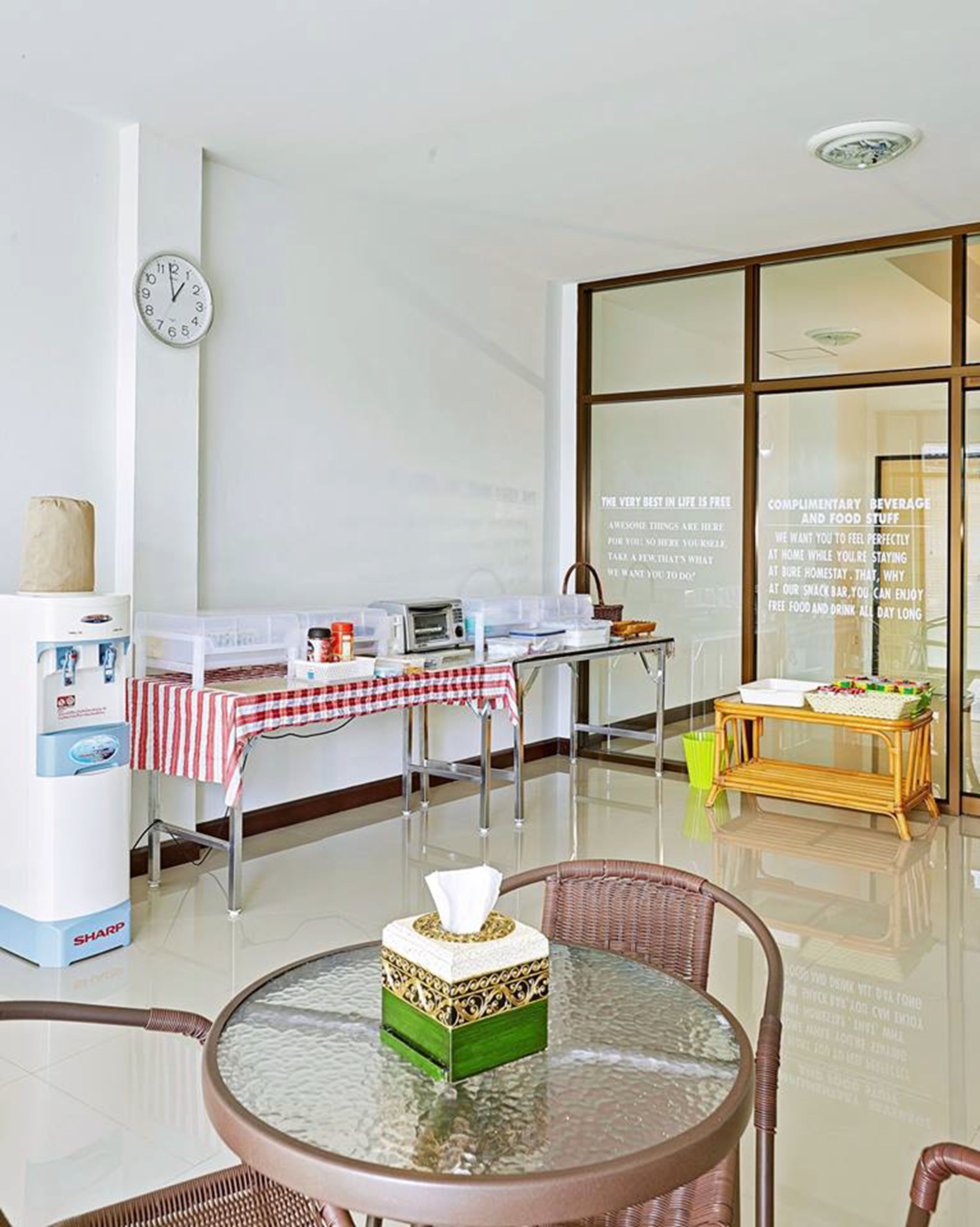 Une chambre moderne avec une table en verre, un fauteuil en osier, un distributeur d’eau Sharp, une horloge et un mur en verre orné d’une citation inspirante.