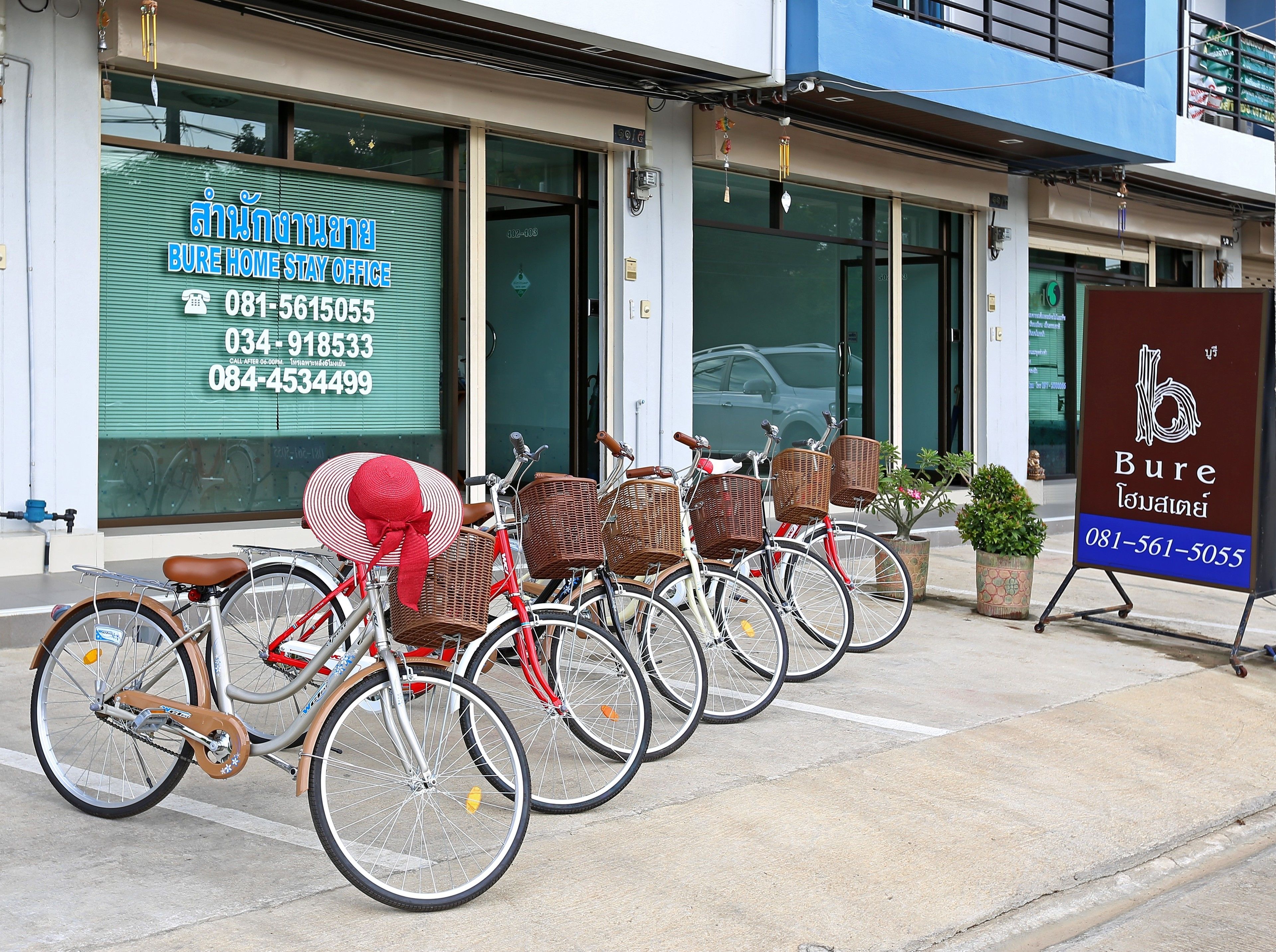 A row of bicycles parked outside a building with a green shutter and a signboard displaying contact information.