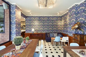 Cozy dining area with a wooden table, sofa, and rug, surrounded by floral wallpaper and a chandelier.