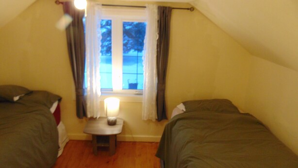 A small, dimly lit room with two beds, a small table, and a window with curtains. - Just You Vacation Home (Gilbert Plains)
