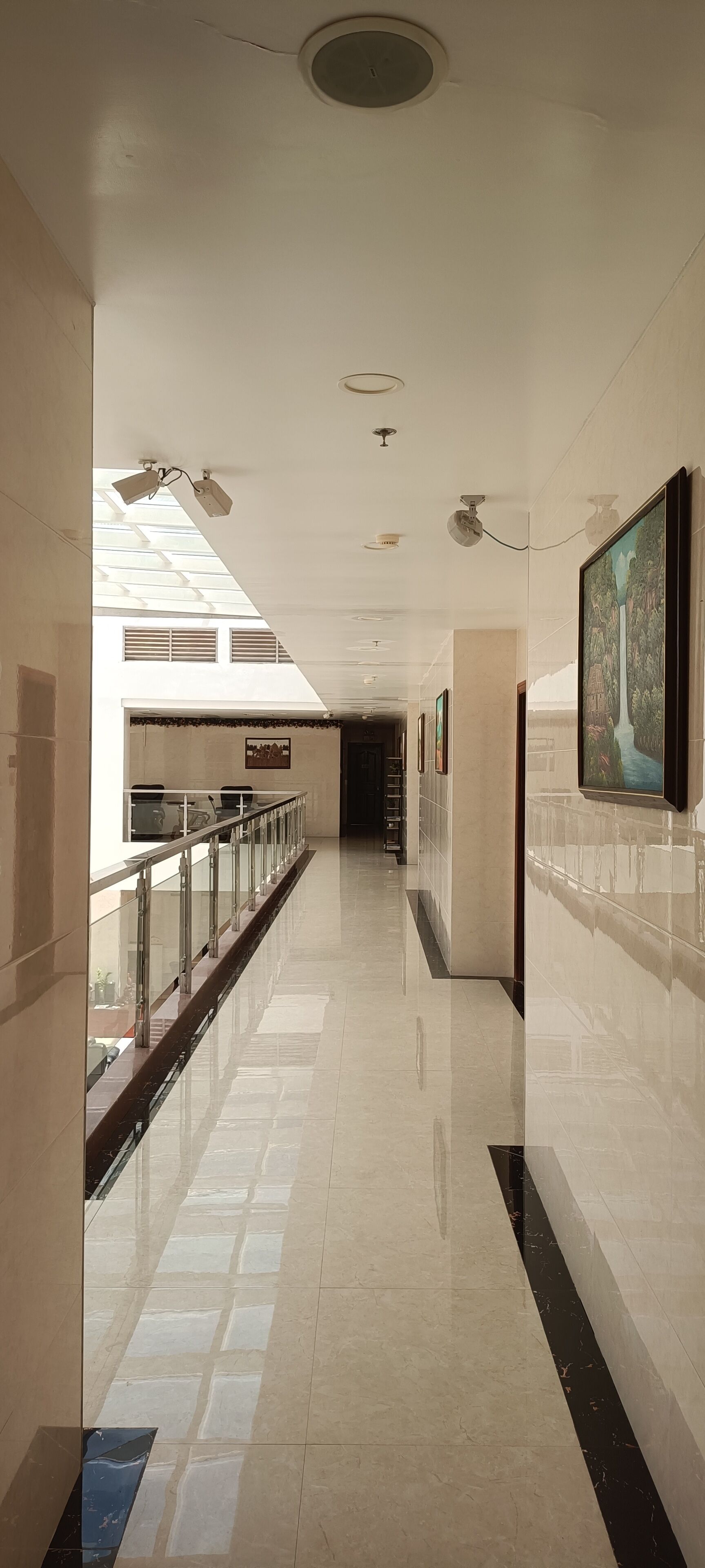 A long, well-lit corridor with a glass railing, tiled floors, and ceiling-mounted lights.