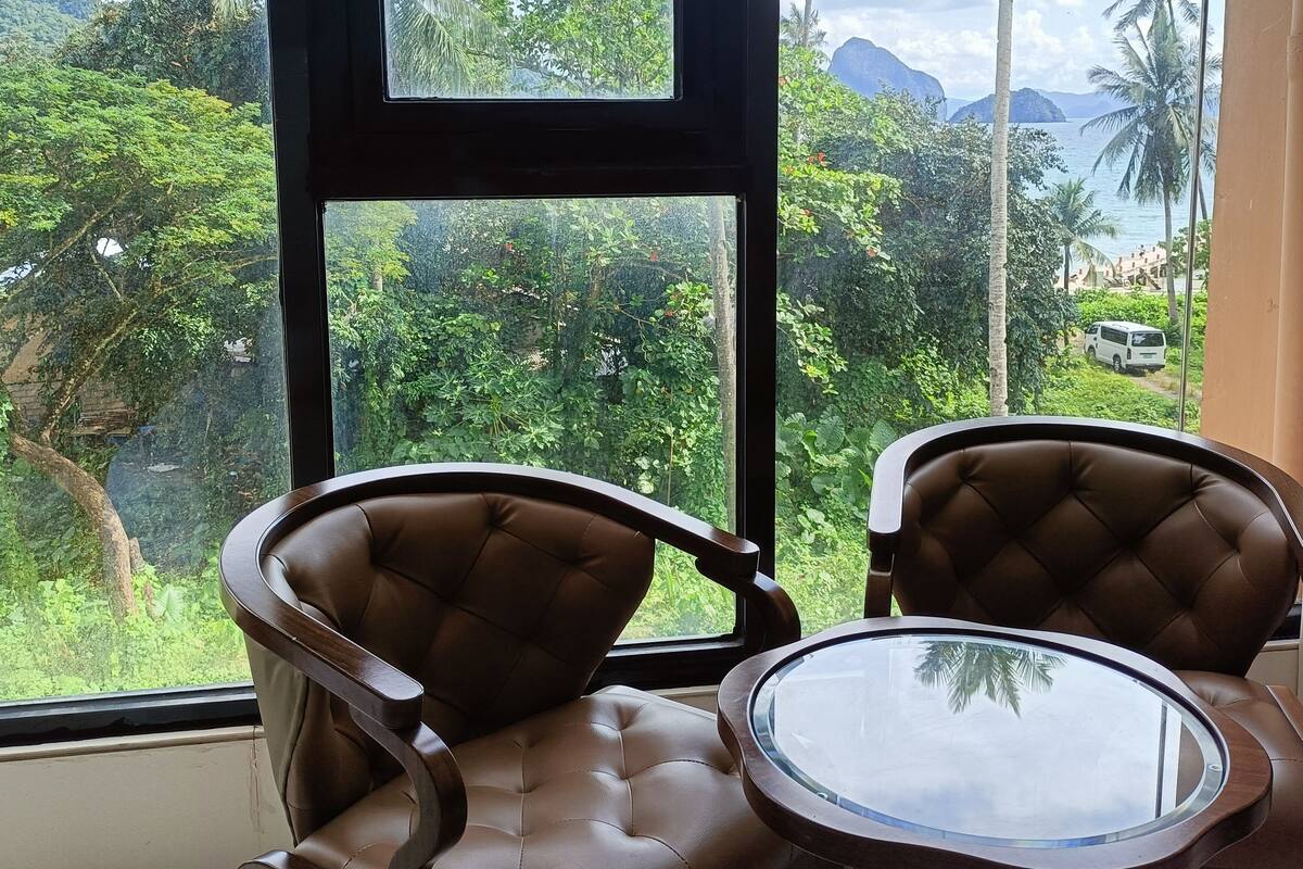 Quarto com duas poltronas de couro marrom e uma pequena mesa redonda, uma janela grande com vista para vegetação exuberante e um corpo d'água.