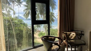 A room with a large window overlooking a beach and ocean, a bed, a chair, and a small table. - One El Nido Suite (El Nido)