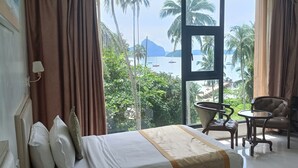 A hotel room with a bed, a chair, a small table, and a view of the sea and palm trees. - One El Nido Suite (El Nido)