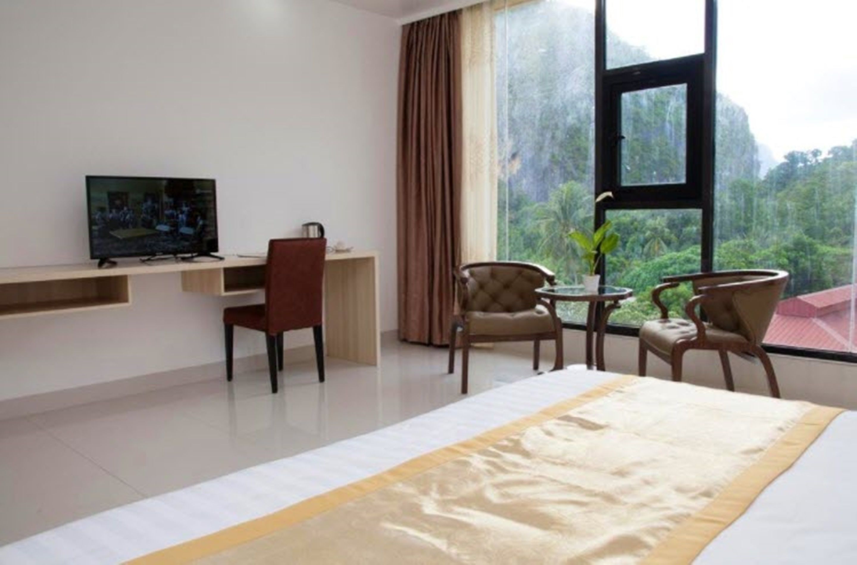 Een hotelkamer met een bed, een televisie op een standaard, een bureau met stoel, een kleine tafel met stoelen en een groot raam met uitzicht op een bos.
