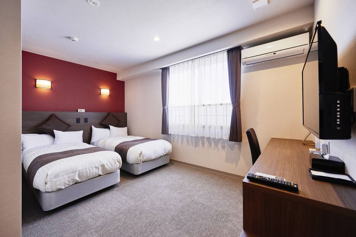 A hotel room with two beds, a desk, a TV, and a window with curtains.