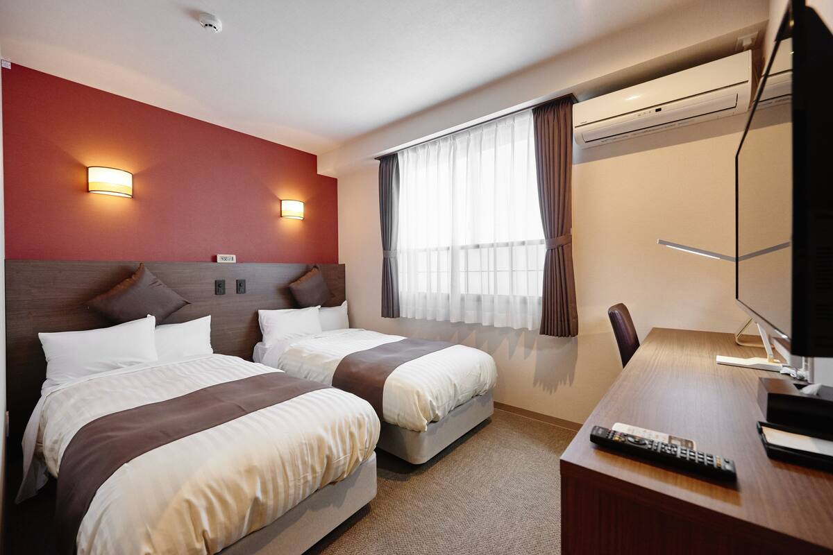 A hotel room with two beds, a desk, a chair, a TV, and an air conditioning unit.
