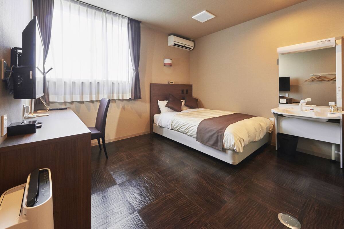 A hotel room with a bed, a desk with a computer, a chair, a desk lamp, an air conditioning unit, a mirror, and a window with curtains.