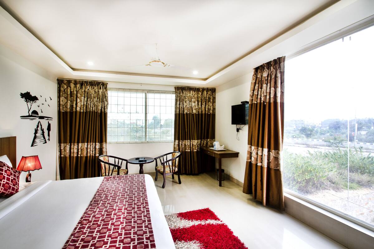 Habitación de hotel con cama, escritorio, dos sillas, ventana con cortinas y vistas al exterior.