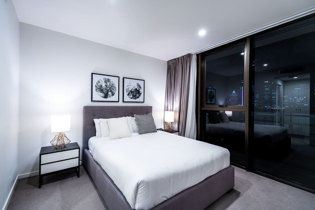 Una habitación de hotel moderna con una cama grande, dos mesitas de noche con lámparas, vistas de la ciudad a través de ventanales del suelo al techo y dos cuadros en la pared.