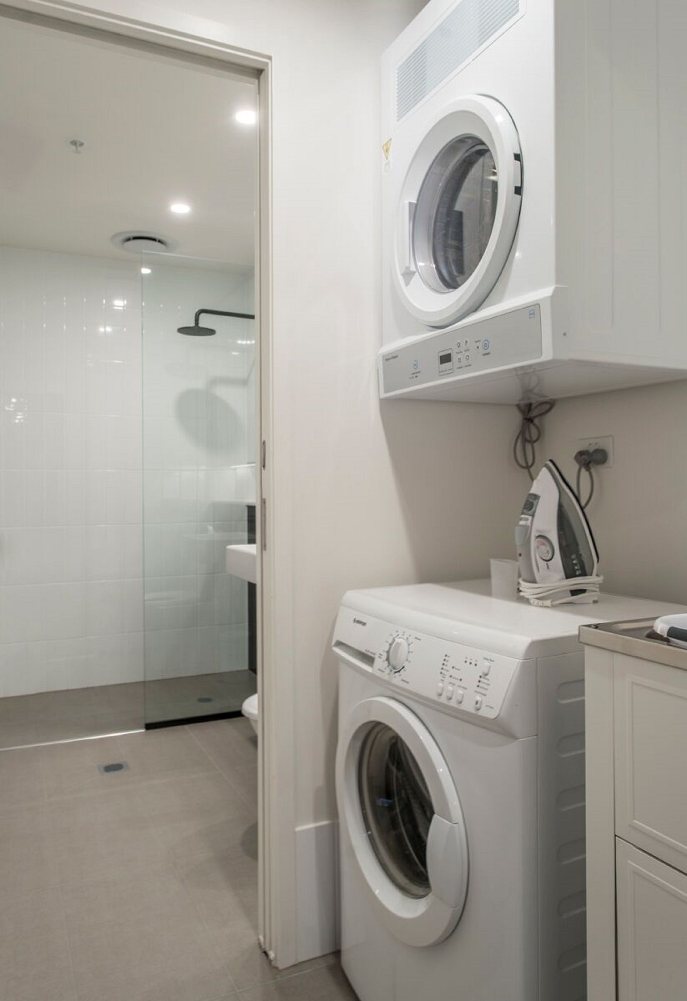 Un espace buanderie compact comprenant un lave-linge et un sèche-linge à chargement frontal, un petit coin repassage et une douche fermée par des parois en verre.