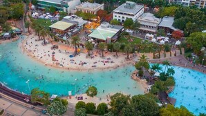 An aerial view of a coastal resort with a central beach area, multiple pools, and surrounding buildings.