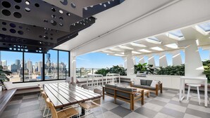 A modern rooftop lounge with a panoramic city view, a wooden dining table, and comfortable seating areas.