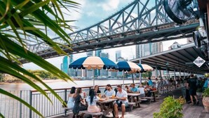 A riverside dining area with outdoor seating under umbrellas, overlooking a bridge and city skyline.