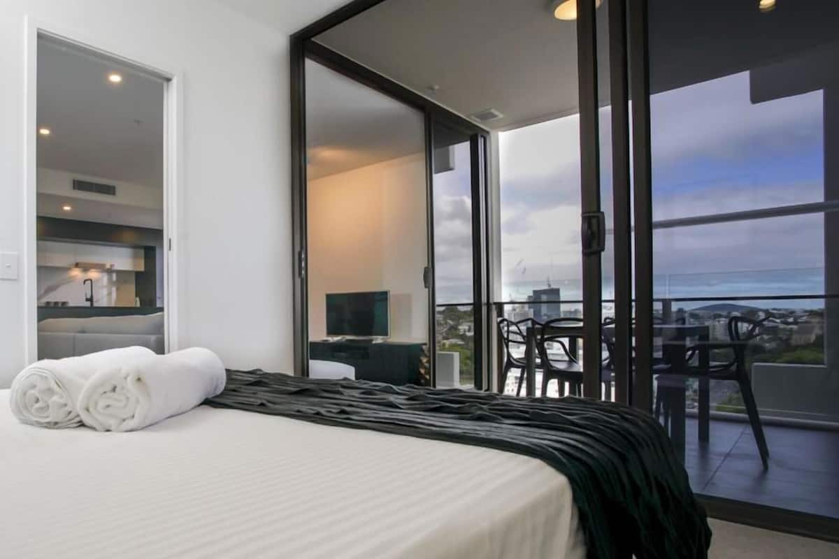 Habitación de hotel con cama, televisor, balcón con sillas y vista a la ciudad.