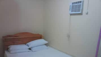 A small, single-bed room with a wooden headboard, white bedding, and a wall-mounted air conditioning unit.