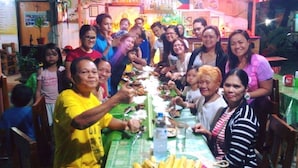 A group of people gathered around a long table with food, in a room with a green tablecloth and various items on the walls. - Emok's Guest House - Hostel (Moalboal)