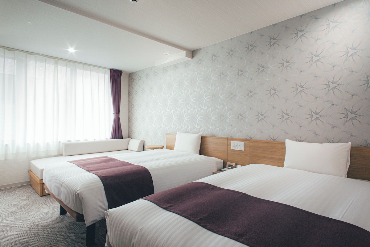A hotel room with two beds, a large window with sheer curtains, a patterned wall, and a wooden headboard.