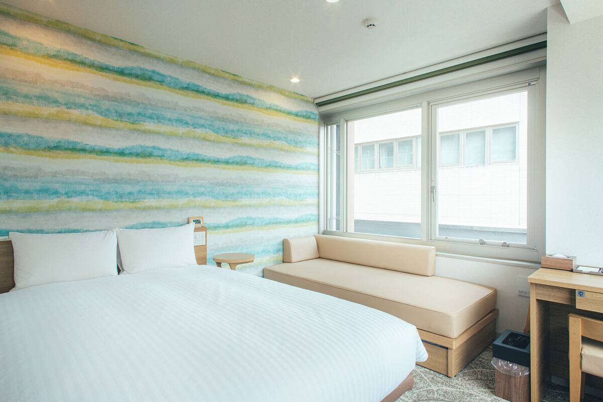 A hotel room with a large bed, a sofa, a small table, and a window with blinds.
