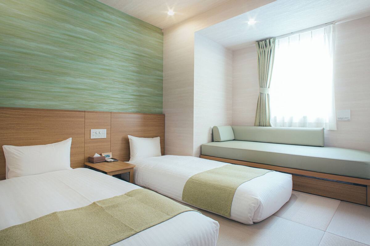A hotel room with two beds, a bench, a nightstand, and a window with curtains.
