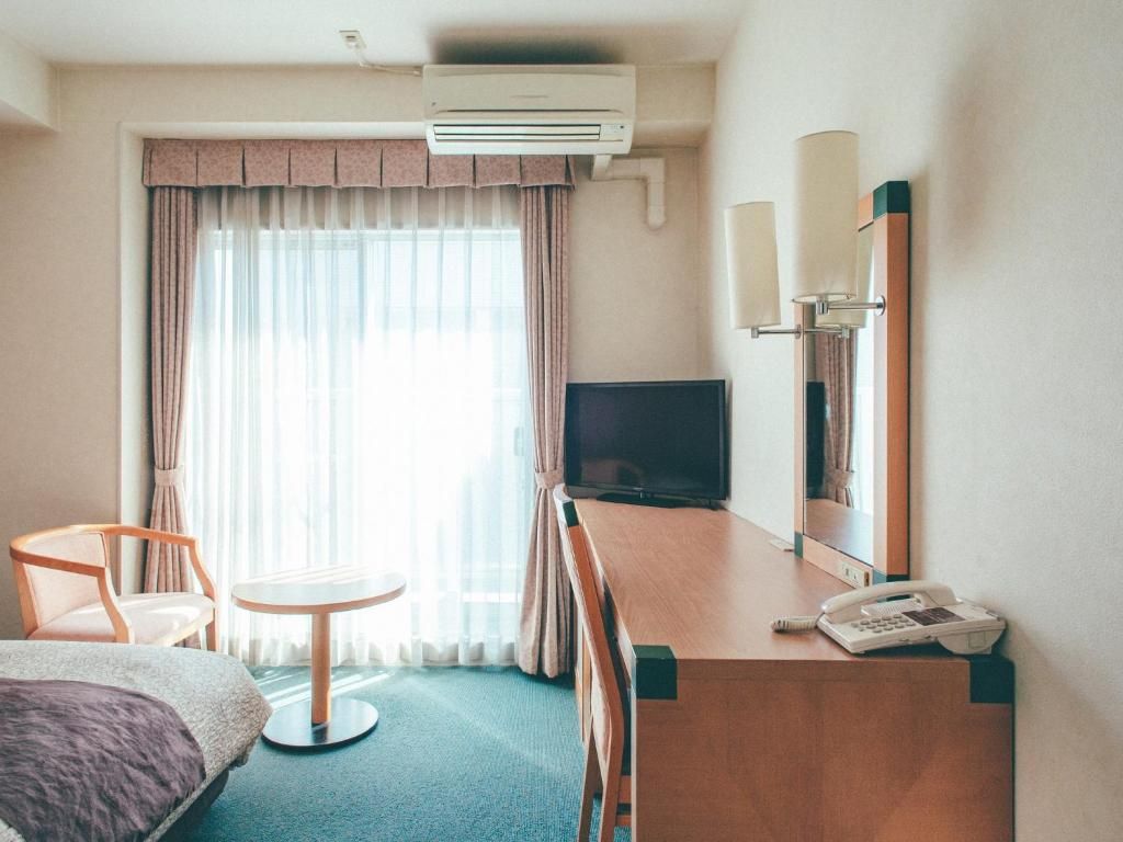A hotel room with a bed, a desk with a telephone, a television, and a window with curtains.
