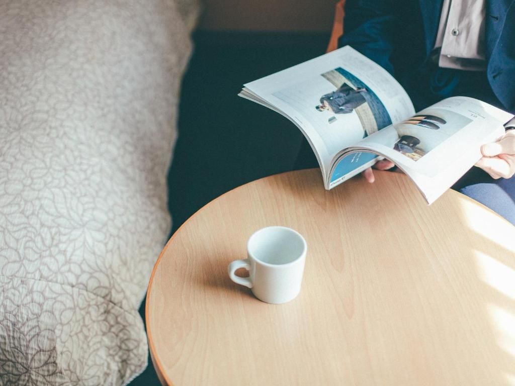 A person reading a magazine on a round wooden table with a white mug beside it.