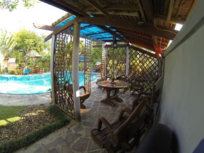 A covered outdoor seating area with stone flooring, wooden furniture, and a pool in the background.