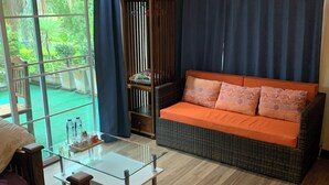 Une pièce avec une table basse en verre, un canapé en osier avec des coussins orange, un cadre de lit en bois et une porte-fenêtre coulissante donnant sur un balcon agrémenté de plantes.