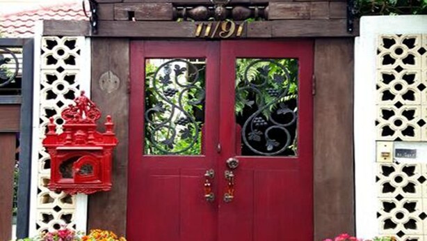A red door with decorative glass panels, a small red lantern, and potted plants on either side. - Villa 91 Guesthouse (Bangkok)