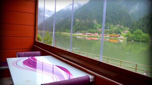 A room with a large window overlooking a lake and mountains, featuring a table and chairs. - Osmanli Suite (Çaykara)